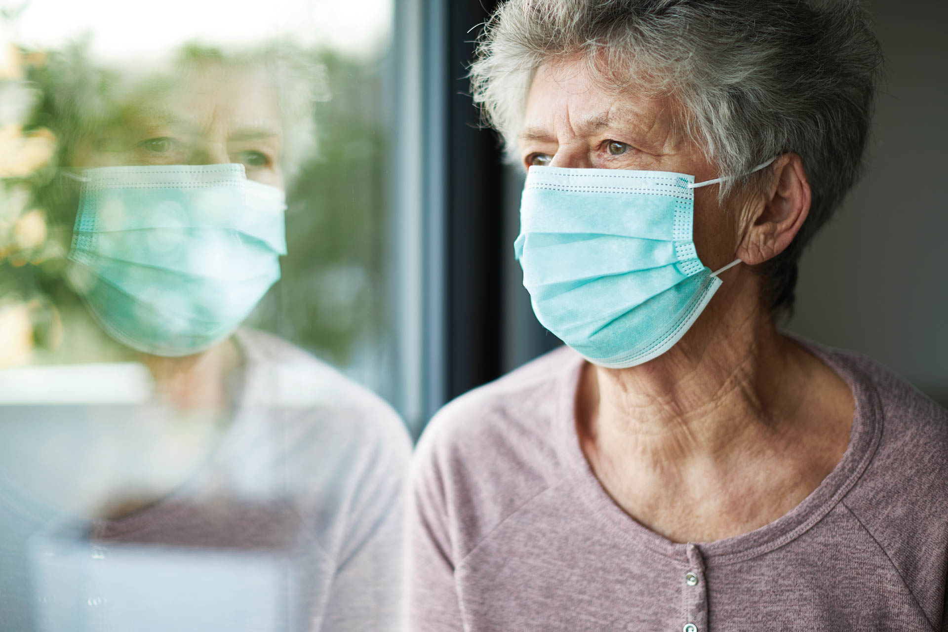 a old woman or grandma is wearing a respirator or surgical mask and is looking out the window while she is in quarantine because of the corona virus.